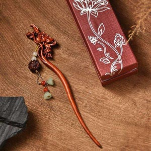 Puede incluir: Un palillo para el cabello de madera tallada con un motivo floral en la parte superior, acentuado con una cadena colgante de pequeñas cuentas y dijes. Al lado hay una caja de regalo rectangular roja decorada con un dibujo lineal blanco de un loto.
