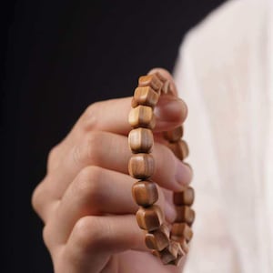 May include: A wooden bracelet with a series of small, square-shaped beads. The beads are a light brown color and appear to be made of wood. The bracelet is held in a person's hand against a dark background.