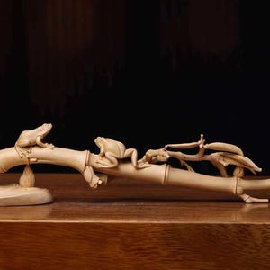 May include: A light beige carved wooden sculpture depicting a horizontal bamboo stalk with three frogs perched along its length. The detailed carving features textured frog skin, segmented bamboo nodes, and small leaves extending from the branch.