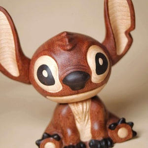 K&ouml;nnte beinhalten: Eine Holzfigur einer Zeichentrickfigur mit gro&szlig;en Ohren, gro&szlig;en Augen und einer dunkelbraunen Nase. Die Figur besteht aus verschiedenen Braunt&ouml;nen, mit helleren Holzakzenten an Ohren und Augen. Die Figur sitzt.