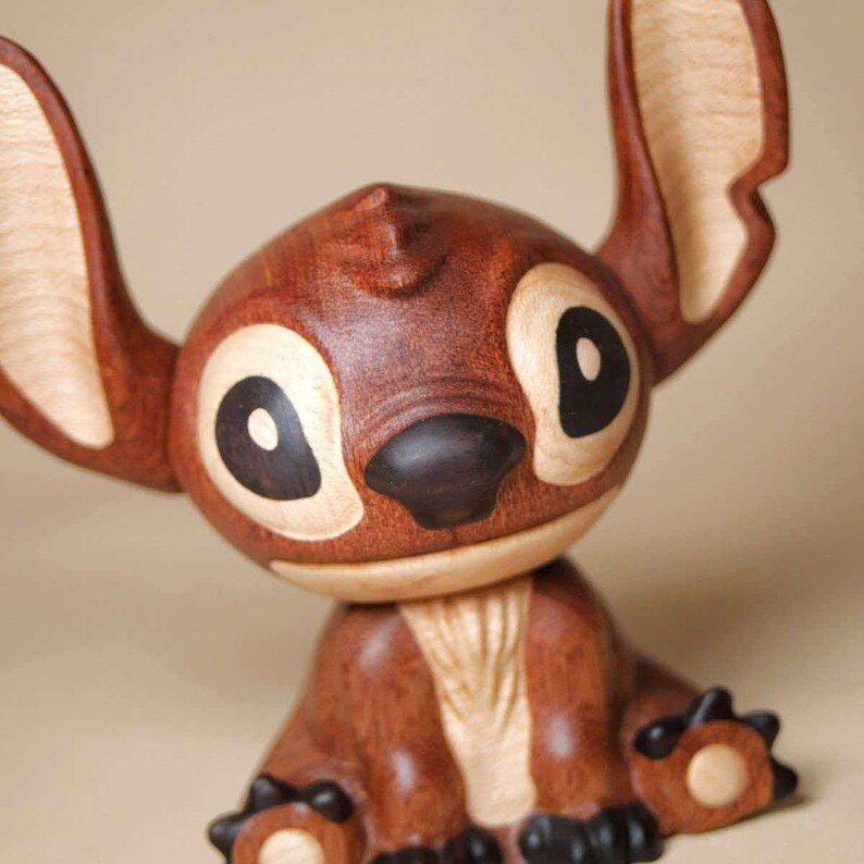 K&ouml;nnte beinhalten: Eine Holzfigur einer Zeichentrickfigur mit gro&szlig;en Ohren, gro&szlig;en Augen und einer dunkelbraunen Nase. Die Figur besteht aus verschiedenen Braunt&ouml;nen, mit helleren Holzakzenten an Ohren und Augen. Die Figur sitzt.