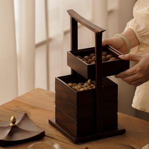May include: A dark brown wooden tiered serving tray with a handle. The tray has three stacked square compartments, two filled with nuts. The handle is held by a person. The tray sits on a wooden table.