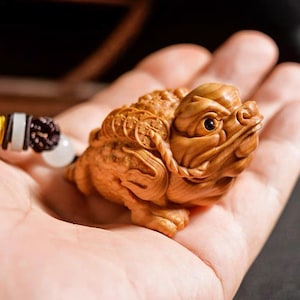 May include: A detailed wooden carving of a toad, featuring intricate textures and a detailed face. The toad is a light brown color and appears to be a small figurine. A beaded necklace is partially visible in the background.