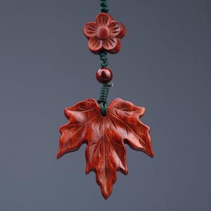 Peut inclure: Un pendentif en bois brun rougeâtre sculpté en forme de feuille d'érable, suspendu par un cordon vert foncé. Au-dessus de la feuille se trouvent une petite perle ronde en bois foncé et une perle sculptée en forme de fleur, reliées par le cordon.