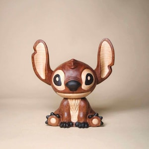K&ouml;nnte beinhalten: Eine Holzfigur der Figur Stitch aus dem Disney-Film Lilo & Stitch. Die braune Figur hat gro&szlig;e Ohren, schwarze Augen und eine schwarze Nase. Die Figur sitzt mit Armen und Beinen nach vorne.