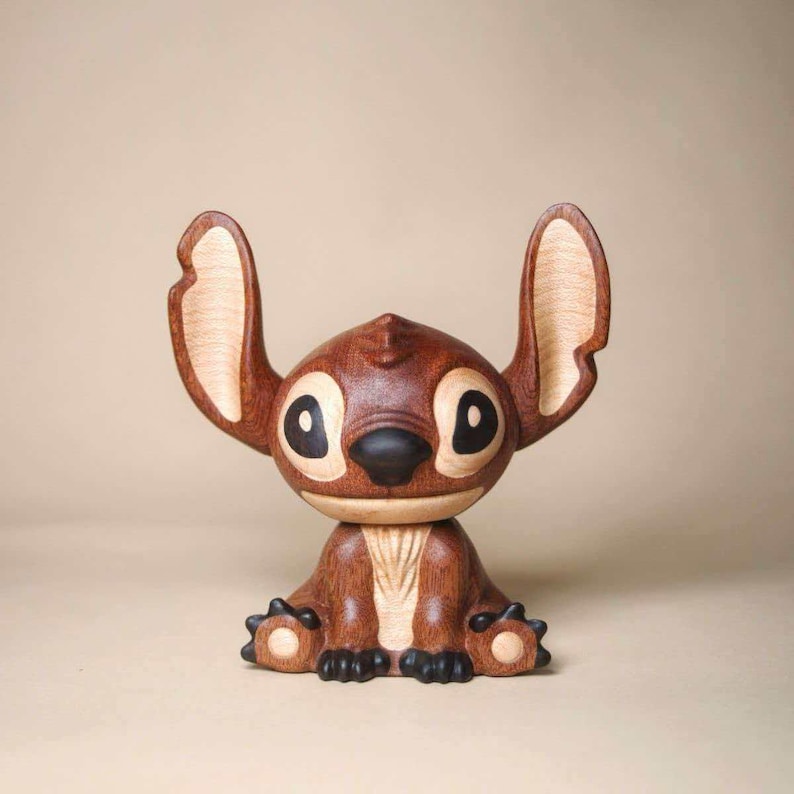 K&ouml;nnte beinhalten: Eine Holzfigur der Figur Stitch aus dem Disney-Film Lilo & Stitch. Die braune Figur hat gro&szlig;e Ohren, schwarze Augen und eine schwarze Nase. Die Figur sitzt mit Armen und Beinen nach vorne.