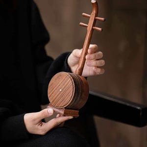 Può includere: Uno strumento musicale cinese in legno, probabilmente un Erhu, con una cassa armonica rotonda e un lungo manico con piroli. Lo strumento è tenuto in mano, mostrando la sua dettagliata venatura del legno e l'artigianato. Uno strumento a corde tradizionale.