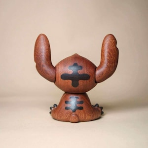 K&ouml;nnte beinhalten: Eine Holzfigur einer Zeichentrickfigur mit gro&szlig;en Ohren und einem runden K&ouml;rper. Die Figur besteht aus dunkelbraunem Holz mit schwarzen Markierungen auf Kopf und K&ouml;rper. Die Ohren sind nach oben gebogen.
