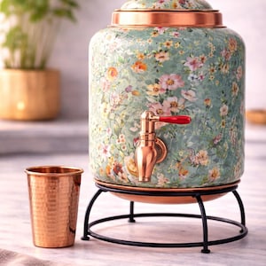 May include: A floral-patterned water dispenser with a copper tap and lid, set on a black metal stand. A copper cup sits beside it. The dispenser has a light green background with colorful flowers.