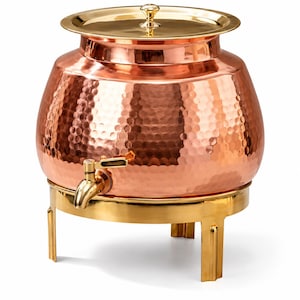 Puede incluir: Un dispensador de bebidas de cobre martillado con una tapa dorada pulida y un grifo dorado a juego. El recipiente redondo descansa sobre un soporte de metal dorado resistente con tres patas, con una abertura ancha en la parte superior.