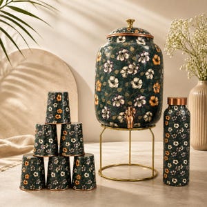 Puede incluir: Un dispensador de bebidas verde oscuro con estampado floral, grifo metálico y tapa a juego, sobre un soporte de alambre dorado. Se acompaña de seis vasos a juego y una botella de agua alta, todos con el mismo estampado floral.