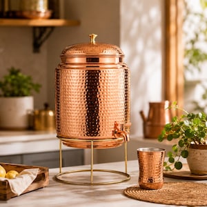Könnte beinhalten: Ein zylindrischer Wasserspender aus Kupfer mit gehämmertem Muster, abnehmbarem Deckel mit kleinem Knauf und einem Metallhahn. Der Spender steht auf einem dünnen, goldfarbenen Drahtgestell. Ein passender, gehämmerter Kupferbecher steht daneben.