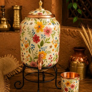 May include: A cream-colored beverage dispenser with a floral pattern in yellow, pink, and blue, set on a black metal stand. A matching cup sits nearby. The dispenser has a copper spigot and a gold-colored lid.