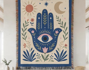 May include: A woven throw blanket featuring a central dark blue hamsa hand with an eye symbol, surrounded by botanical illustrations, a sun, and a crescent moon on a cream background. The blanket has a dark blue border and fringed edges.