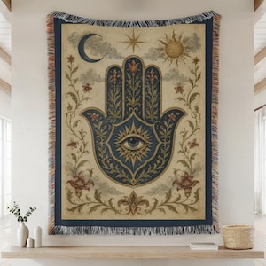 May include: A woven throw blanket featuring a central dark blue hamsa hand symbol with an eye, surrounded by floral vines, a crescent moon, a sun, and a star. The design is set against a cream background with a dark blue border and fringed edges.