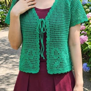 May include: A bright green crochet cardigan featuring short sleeves, a scalloped edge trim, and a front tie closure. The garment is crafted with an open-work grid pattern and is worn over a solid dark red sleeveless top and a matching red skirt.