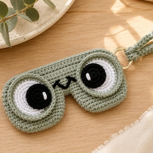 May include: A sage green crocheted sleep mask featuring two large circular eyes with white and black yarn details, a small black stitched smile, and a matching braided wrist strap attached by a gold-toned metal ring.