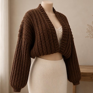 May include: A dark brown, long-sleeved, cropped cardigan featuring a thick, vertical ribbed knit texture. The garment has a wide collar, dropped shoulders, and cuffed sleeves, worn over a cream-colored, textured, sleeveless top and a matching skirt.