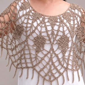 Peut inclure: Un poncho en crochet beige présentant un motif complexe en toile ajourée avec des motifs floraux solides intégrés dans le design. Le vêtement possède une large encolure ronde et un ourlet à franges qui tombe librement sur les épaules.