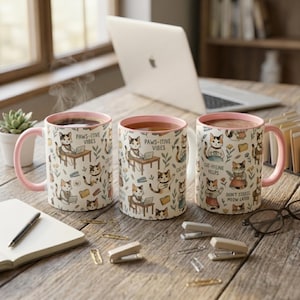 Puede incluir: Tres tazas de cerámica con asas y bordes rosas, con un diseño de gatos y las frases "PAWS-ITIVE VIBES", "CAT-FFEINE HELPS" y "DON'T STEESS MEOW LATER". Las tazas contienen una bebida caliente y están sobre una superficie de madera.