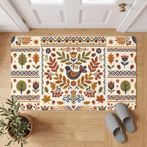 May include: A rectangular doormat featuring a cream background with a repeating pattern of birds, flowers, leaves, and acorns in shades of orange, blue, green, and brown. The design includes geometric borders with zigzags and diamonds.