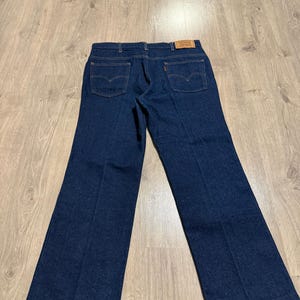 May include: Dark blue denim jeans with a classic five-pocket design. The jeans feature a straight leg cut and a small red tag on the back pocket. The jeans are laid flat on a wooden surface.