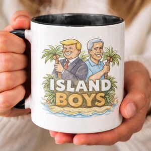 Op de afbeelding: Witte keramische mok met een zwarte binnenkant en handvat. De mok heeft een cartoonillustratie van twee mannen die palmbomen vasthouden, met de tekst "ISLAND BOYS" in een tropisch ontwerp.