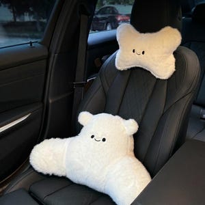 May include: Two white plush car seat pillows with smiling faces. One is a bone-shaped headrest pillow, and the other is a lumbar support pillow. The pillows are on a black leather car seat.