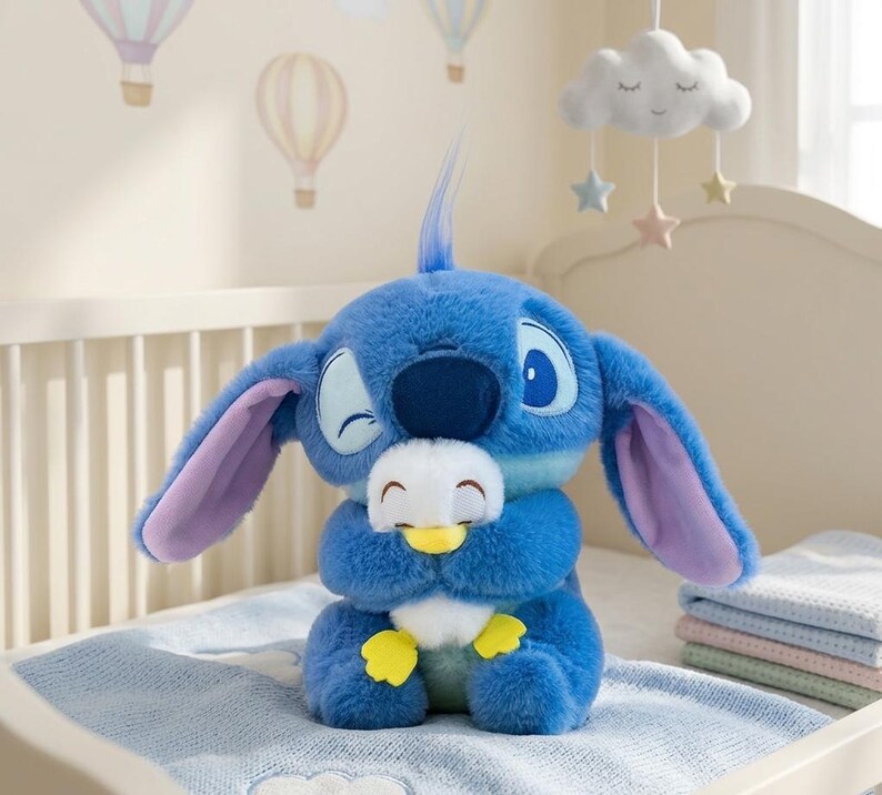 May include: A blue plush toy of a cartoon character with large ears and a winking expression, holding a smaller white and yellow plush toy. The plush toys are on a light blue blanket, with a crib and decorative hanging mobile in the background.