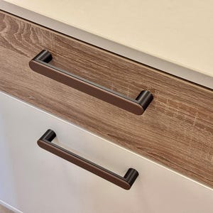 Puede incluir: Primer plano de un mueble con dos cajones. El cajón superior es de laminado con vetas de madera y el cajón inferior es blanco. Cada cajón tiene un tirador rectangular marrón oscuro. La parte superior del mueble es de color crema claro.