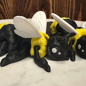 May include: A handcrafted bee ornament. The bee has a black body with yellow stripes, white wings, and a smiling face. The figurine is lying down, with its legs extended. The bee is a decorative item.