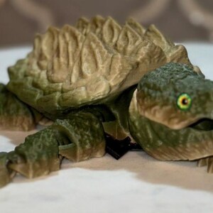 May include: A 3D printed articulated alligator snapping turtle figurine featuring a textured olive green body and a tan, ridged shell. The turtle has a detailed head with a yellow and black eye, a wide mouth, and segmented limbs that allow for movement.