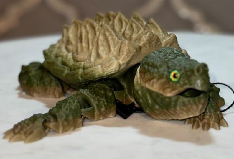 May include: A 3D printed articulated alligator snapping turtle figurine featuring a textured olive green body and a tan, ridged shell. The turtle has a detailed head with a yellow and black eye, a wide mouth, and segmented limbs that allow for movement.