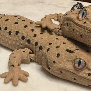 May include: Two tan gecko figurines with black spots and blue and pink eyes. One gecko is lying down, and the other is on top. The geckos have detailed skin textures and are posed on a white surface.