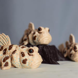 May include: A set of three ceramic mugs with a speckled, matte finish. The mugs are in shades of cream, light gray, and dark gray. Each mug has a simple, minimalist design and a rounded handle.