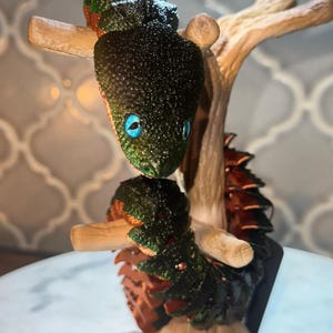May include: A small, articulated creature figurine with a textured, dark green and metallic copper body, featuring bright blue eyes. The creature is coiled around a light brown, branch-like stand, showcasing a segmented, scaly appearance.