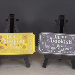 May include: Two decorative ticket-shaped signs, one yellow and one gray, each displayed on a small easel. The signs read "In my bookish era BOOK CLUB LIFETIME MEMBERSHIP" with decorative elements. The yellow sign has purple accents, and the gray sign has lavender accents.