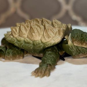 May include: A 3D printed articulated snapping turtle figurine featuring a textured, tan-colored shell with prominent ridges and a dark green head, limbs, and tail. The turtle has bright yellow eyes with black pupils and a detailed, scaly skin texture.