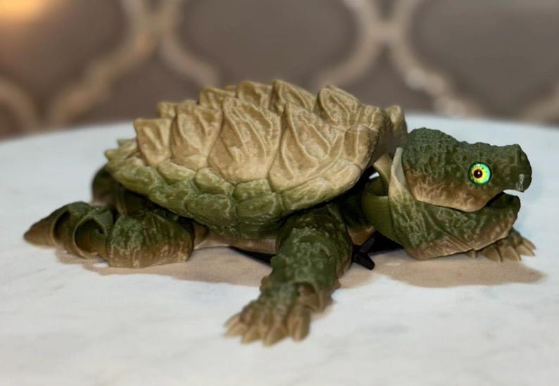 May include: A 3D printed articulated snapping turtle figurine featuring a textured, tan-colored shell with prominent ridges and a dark green head, limbs, and tail. The turtle has bright yellow eyes with black pupils and a detailed, scaly skin texture.