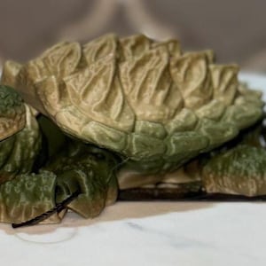 May include: An articulated snapping turtle figurine featuring a textured, olive-green head and limbs with a prominent, spiked, tan and green shell. The creature has a detailed, rugged appearance with visible joints on the legs and a realistic eye.