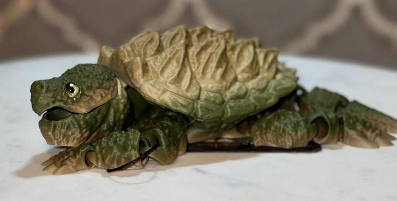 May include: An articulated snapping turtle figurine featuring a textured, olive-green head and limbs with a prominent, spiked, tan and green shell. The creature has a detailed, rugged appearance with visible joints on the legs and a realistic eye.