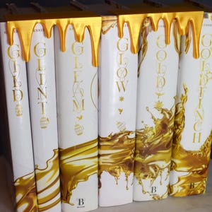 May include: A collection of hardcover books with white covers and gold lettering. The titles include "Gild," "Glint," "Gleam," "Glow," "Goldfinch." A gold-colored decorative element resembling dripping paint adorns the top of the books. The books are arranged vertically.