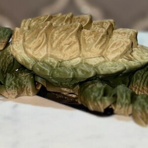 May include: A 3D printed articulated snapping turtle figurine featuring a textured olive green body and a tan, ridged shell. The creature has a detailed head with visible eyes, a wide mouth, and segmented limbs that allow for movement and posing.