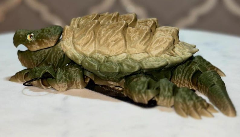 May include: A 3D printed articulated snapping turtle figurine featuring a textured olive green body and a tan, ridged shell. The creature has a detailed head with visible eyes, a wide mouth, and segmented limbs that allow for movement and posing.