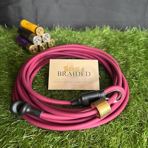 Puede incluir: Una correa enrollada de color burdeos con un asa negra y un cierre color latón. El producto está etiquetado como "Brass & Braided" e incluye casquillos de cartuchos de escopeta. La correa descansa sobre césped verde artificial.