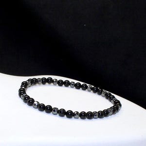 Puede incluir: Una pulsera de cuentas negras con cuentas mate y metálicas alternas. La pulsera se exhibe sobre una superficie blanca sobre un fondo negro. Las cuentas tienen aproximadamente 0,6 cm de diámetro.