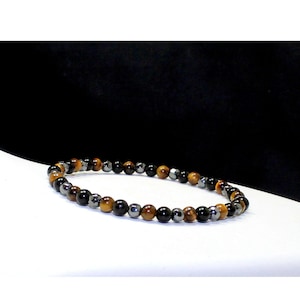 May include: A beaded bracelet featuring a repeating pattern of round, polished tiger eye beads in shades of golden brown, smooth black beads, and faceted metallic gray beads arranged in a circular loop on a white surface.