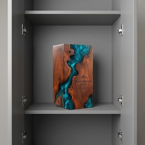 May include: A rectangular wooden cremation urn featuring a central river-like inlay of translucent teal epoxy resin. The front surface is engraved with the text Robert Thompson, the dates 1940-2023, and the phrase Forever in our hearts.