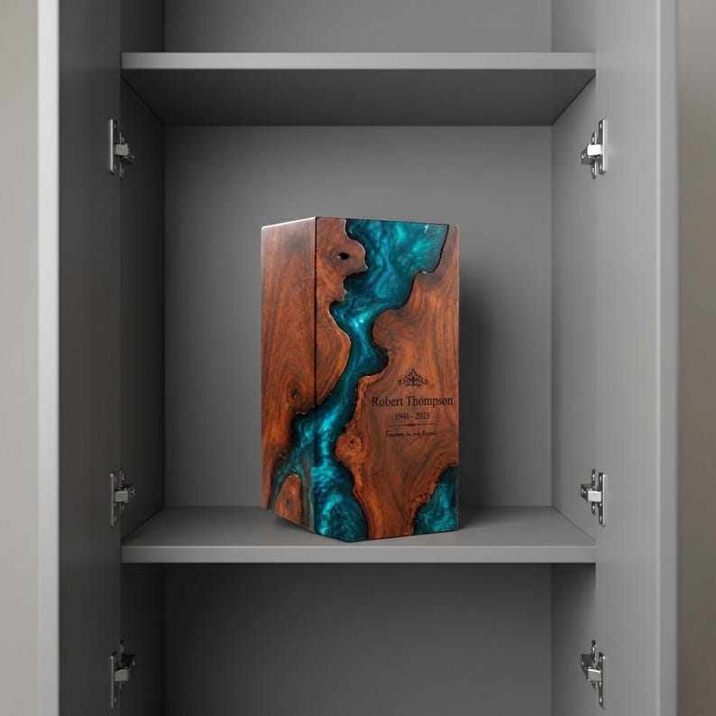 May include: A rectangular wooden cremation urn featuring a central river-like inlay of translucent teal epoxy resin. The front surface is engraved with the text Robert Thompson, the dates 1940-2023, and the phrase Forever in our hearts.