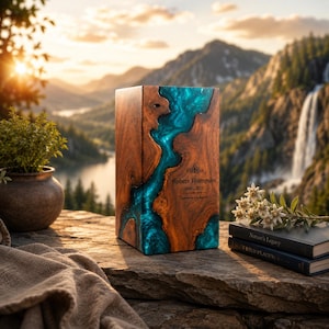 May include: A rectangular wooden cremation urn featuring a central river-like inlay of swirling turquoise resin. The front surface is engraved with the name Robert Thompson, the years 1948-2023, and a small decorative flourish above the text.